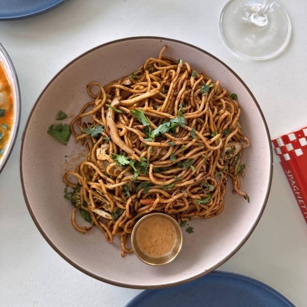 A plate of brown noodles with green herbs and a small cup of sauce on a white table.