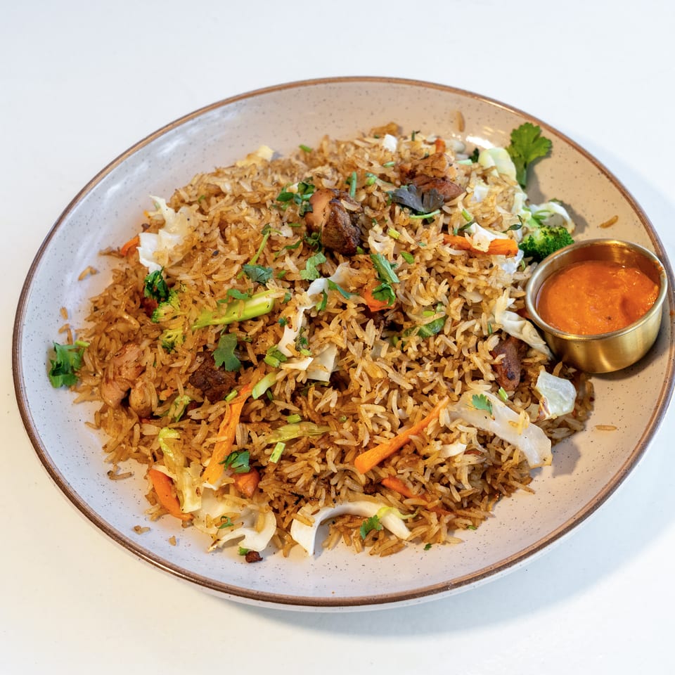Plate of spiced rice with chicken, vegetables, herbs, and a small bowl of orange dipping sauce.