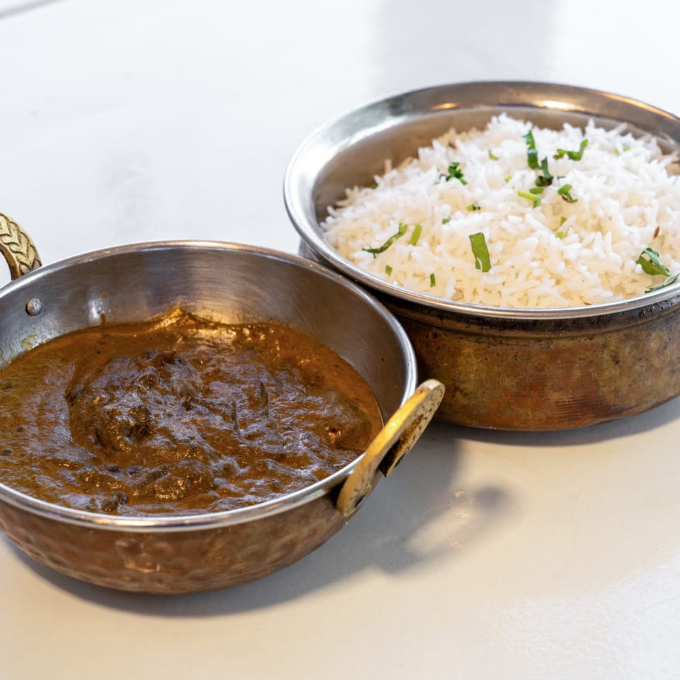 Indian curry and garnished basmati rice served in ornate metal bowls on a white tabletop.