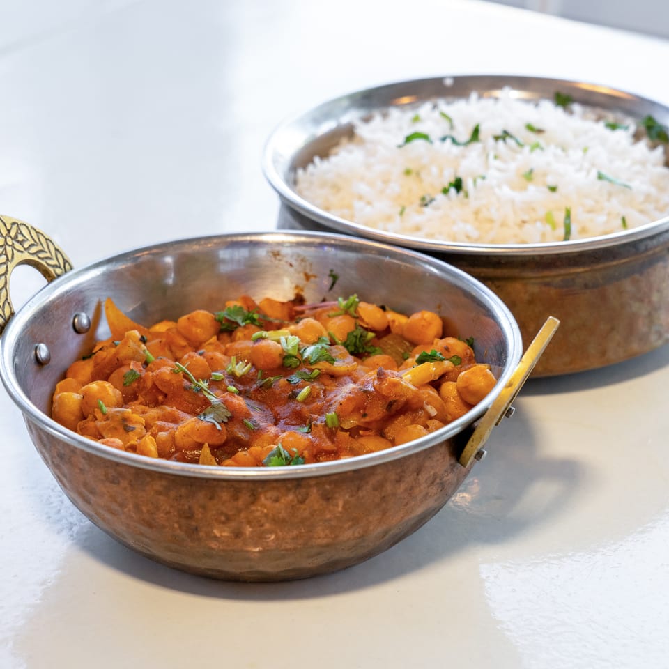 Two metal serving dishes hold chickpea curry garnished with cilantro and white rice sprinkled with herbs.
