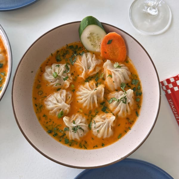 A bowl of momos in a rich orange sauce, garnished with fresh herbs, cucumber, and carrot.