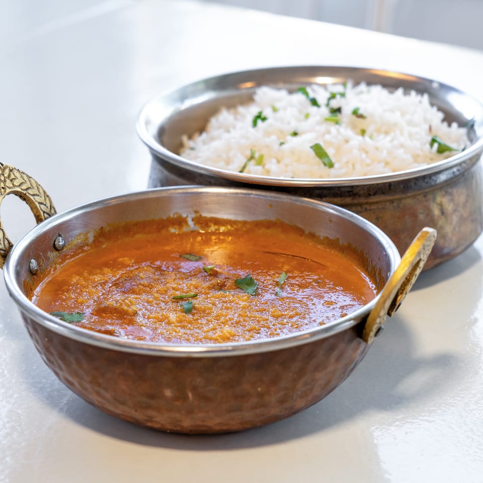 Copper serving bowls hold a garnished red curry and white rice on a bright tabletop.