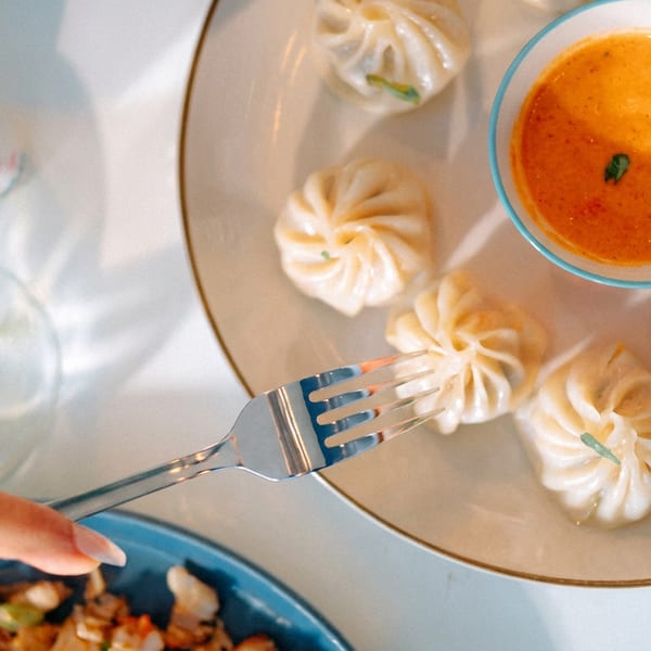 Hand with a fork picking up a steamed dumpling from a plate with orange sauce and wine.