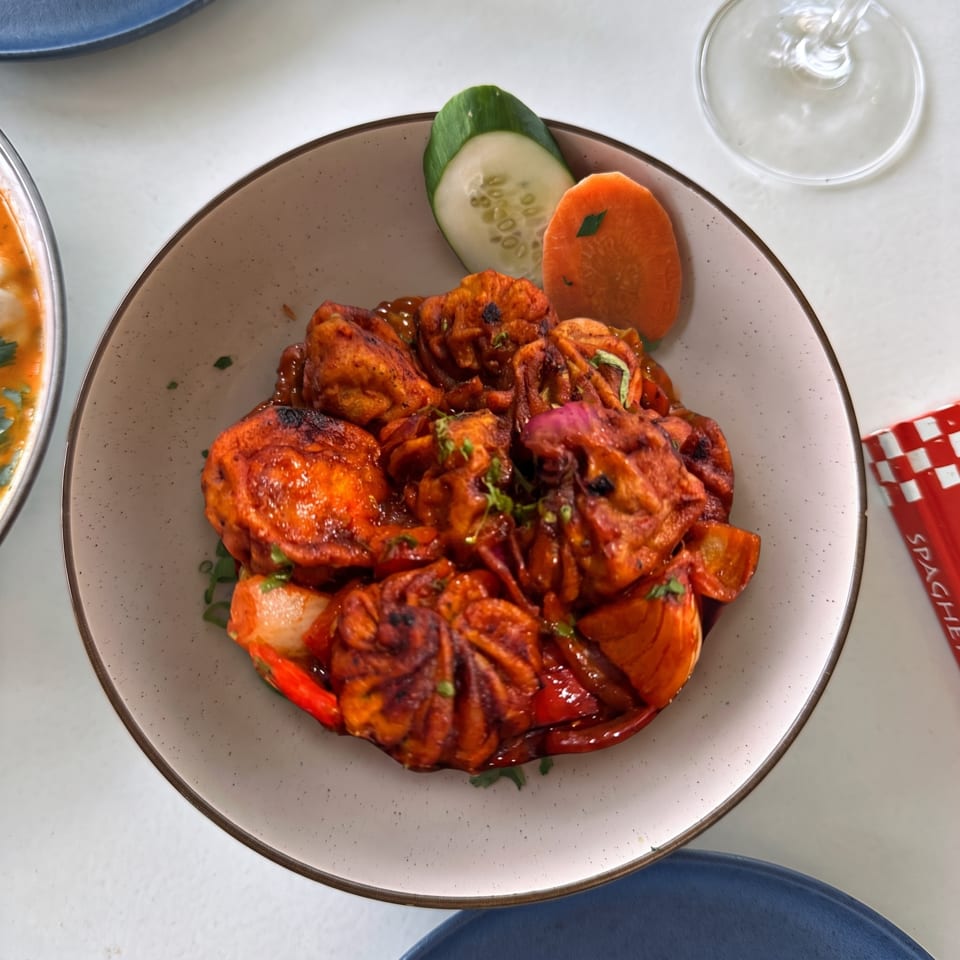 A bowl of spicy red momos with sliced cucumber and carrot on a white table.