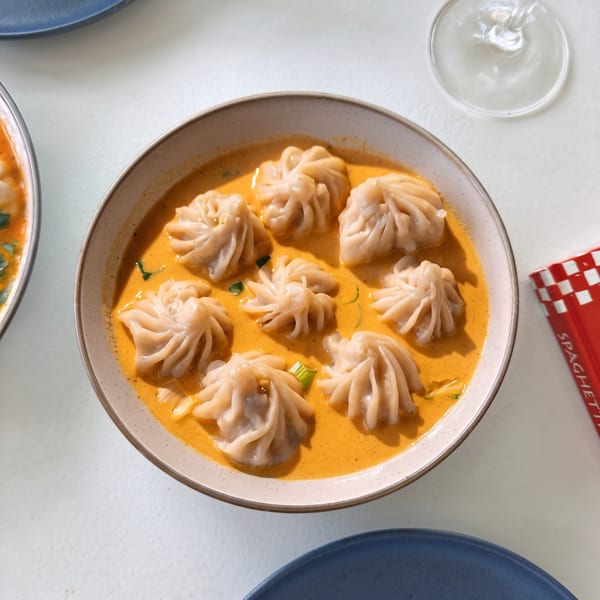 A bowl of momo dumplings in a rich orange sauce, garnished with green herbs.