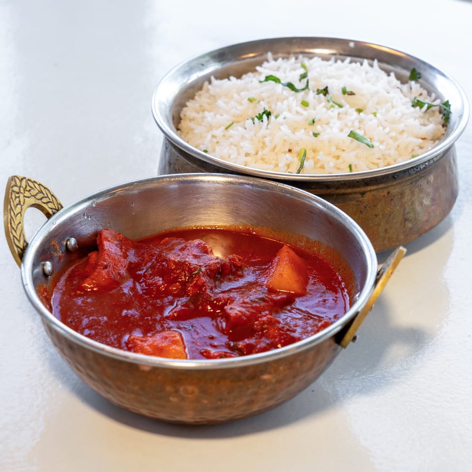 Copper serving bowls hold white herb-garnished rice and red tomato-based curry on a restaurant table.